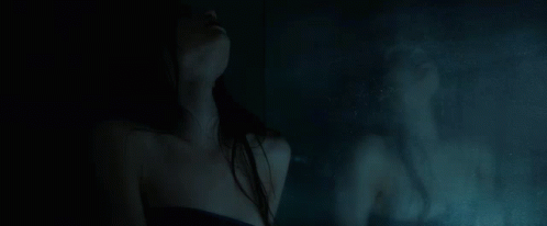 A Woman Is Standing In Front Of A Mirror In A Dark Room With Her Reflection In The Mirror . GIF
