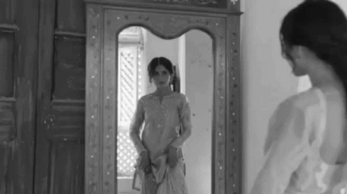 A Woman Is Standing In Front Of A Mirror Looking At Her Reflection . GIF