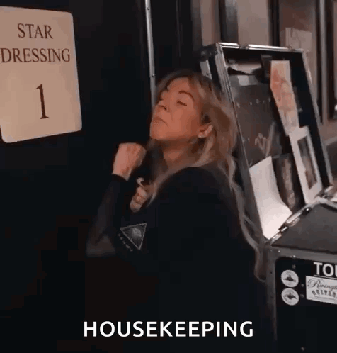 A Woman Is Standing In Front Of A Sign That Says Star Dressing 1 And A Box That Says Housekeeping . GIF