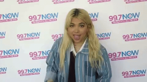 A Woman Is Standing In Front Of A Wall That Says 99.7 Now GIF