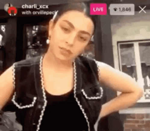 A Woman Is Standing In Front Of A Window Wearing A Black Vest And Earrings . GIF