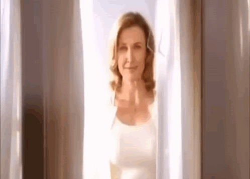 A Woman Is Standing In Front Of A Window With White Curtains And Smiling . GIF