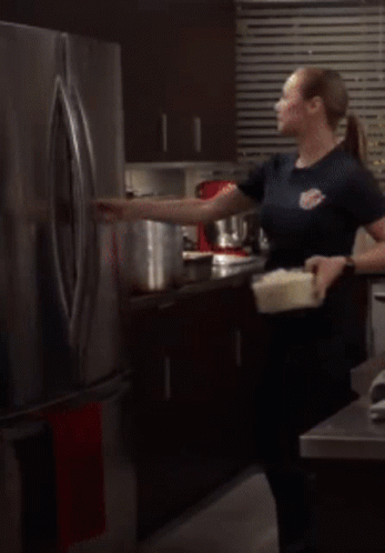 A Woman Is Standing In Front Of An Open Refrigerator Holding A Container GIF