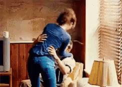 A Woman Is Standing Next To A Man In A Living Room Hugging Him . GIF