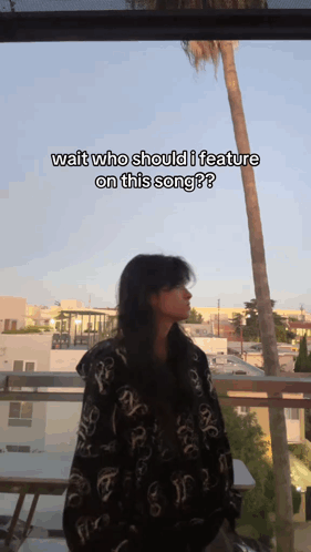 A Woman Is Standing On A Balcony With The Caption Wait Who Should I Feature On This Song ? GIF