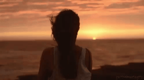 A Woman Is Standing On A Beach Watching The Sunset Over The Ocean . GIF