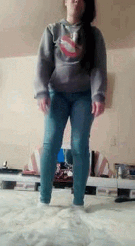 A Woman Is Standing On A Bed Wearing A Hoodie And Jeans . GIF