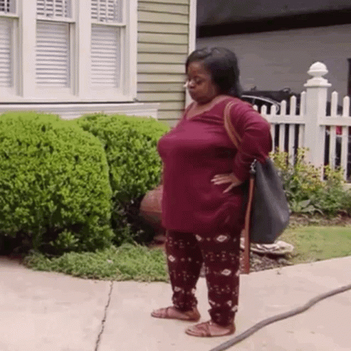 A Woman Is Standing On A Sidewalk In Front Of A House Holding A Hose . GIF