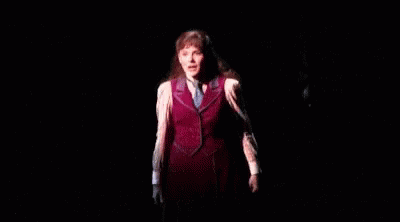 A Woman Is Standing On A Stage In A Dark Room Wearing A Red Vest And Tie . GIF