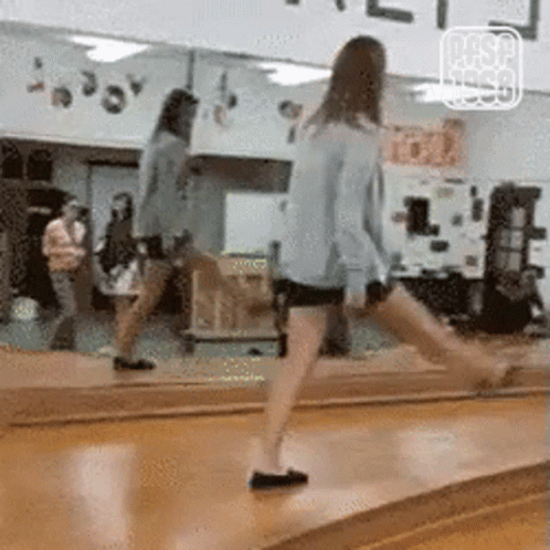 A Woman Is Standing On A Wooden Floor In Front Of A Large Mirror . GIF