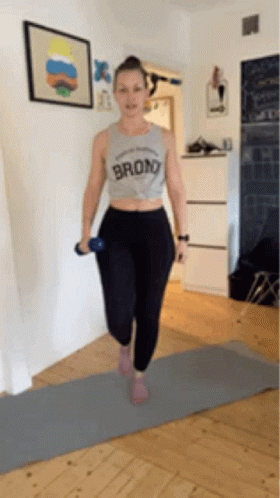 A Woman Is Standing On A Yoga Mat Holding A Dumbbell And Wearing A Shirt That Says Bronx . GIF
