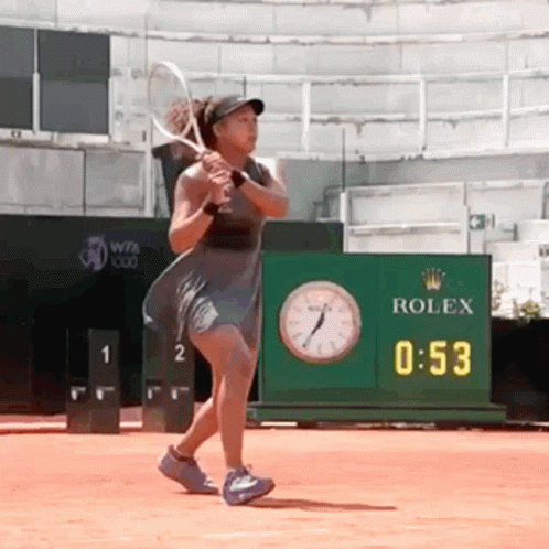 A Woman Is Swinging A Tennis Racquet In Front Of A Rolex Clock GIF