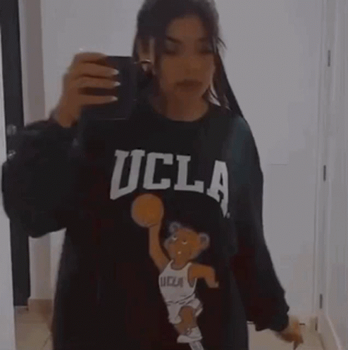 A Woman Is Taking A Picture Of Herself In A Mirror While Wearing A Ucla Sweatshirt . GIF