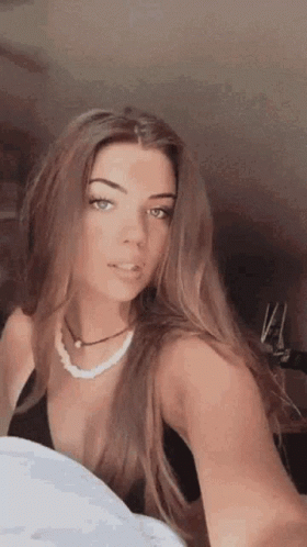 A Woman Is Taking A Selfie With Her Phone While Wearing A Black Top And A Pearl Necklace . GIF