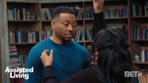 A Woman Is Touching A Man 'S Face In Front Of A Bookshelf With Assisted Living Written On It GIF