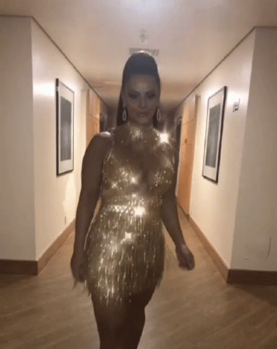 A Woman Is Walking Down A Hallway Wearing A Gold Dress GIF