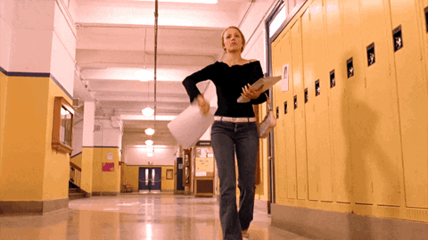 A Woman Is Walking Down A Hallway With Yellow Lockers And A Sign On The Wall That Says ' A ' On It GIF