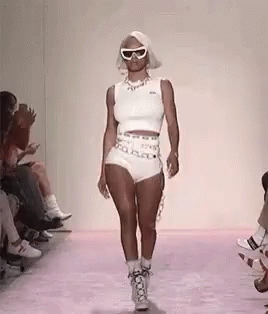 A Woman Is Walking Down A Runway At A Fashion Show Wearing Shorts And Sunglasses . GIF