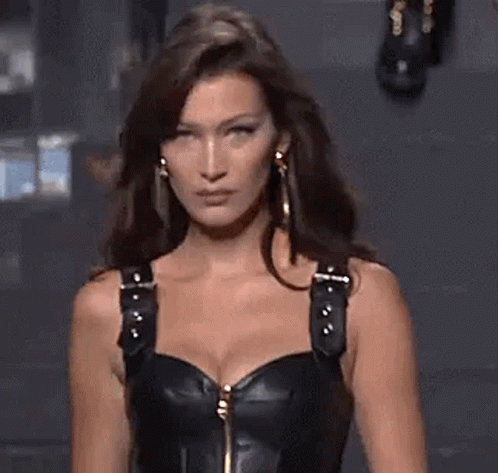 A Woman Is Walking Down A Runway Wearing A Black Leather Corset And Earrings . GIF