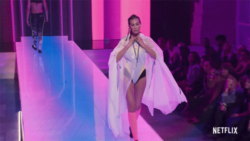 A Woman Is Walking Down A Runway Wearing A White Cape And Pink Socks . GIF