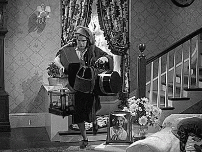 A Woman Is Walking Down A Set Of Stairs In A Living Room Holding A Dog . GIF