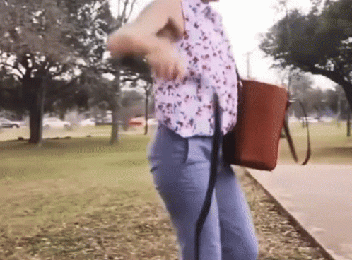 A Woman Is Walking Down A Sidewalk In A Park With A Brown Bag GIF