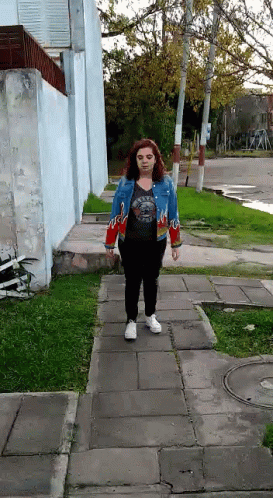 A Woman Is Walking Down A Sidewalk Wearing A Blue Jacket With Flames On The Sleeves GIF