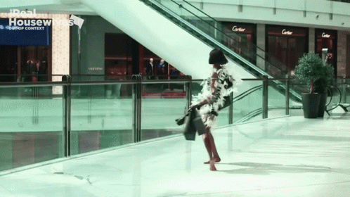 A Woman Is Walking In A Mall With A Sign That Says Real Housewives On It GIF