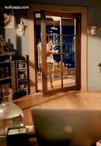 A Woman Is Walking Through A Sliding Glass Door In A Living Room Next To An Apple Laptop . GIF