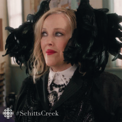 A Woman Is Wearing A Black Feathered Hat With The Hashtag Schitts Creek On The Bottom GIF