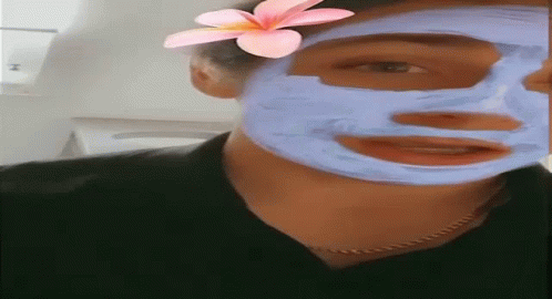 A Woman Is Wearing A Blue Mask On Her Face With A Pink Flower In Her Hair . GIF