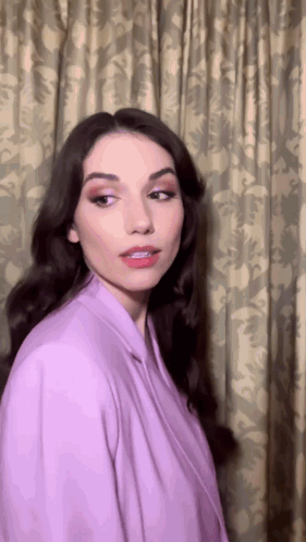 A Woman Is Wearing A Purple Jacket And Standing In Front Of A Curtain GIF