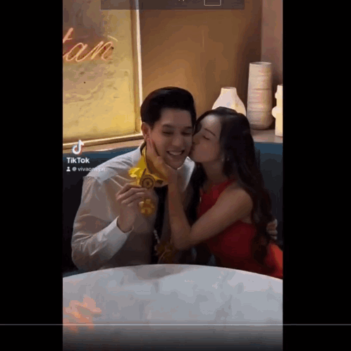 A Woman Kisses A Man On The Cheek In Front Of A Sign That Says " Tan " GIF