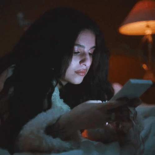 A Woman Laying On A Bed Looking At A Cell Phone GIF