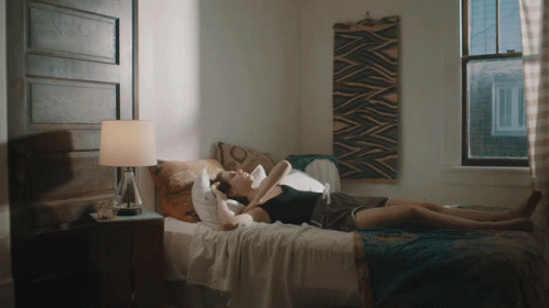 A Woman Laying On A Bed With A Lamp And A Window Behind Her GIF