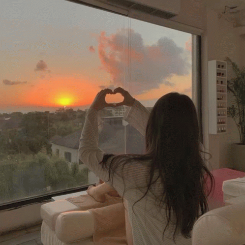 A Woman Making A Heart Shape With Her Hands While Looking Out A Window At A Sunset GIF