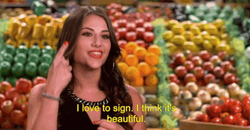 A Woman Says I Love To Sign I Think It 'S Beautiful In Front Of A Bunch Of Fruits And Vegetables GIF