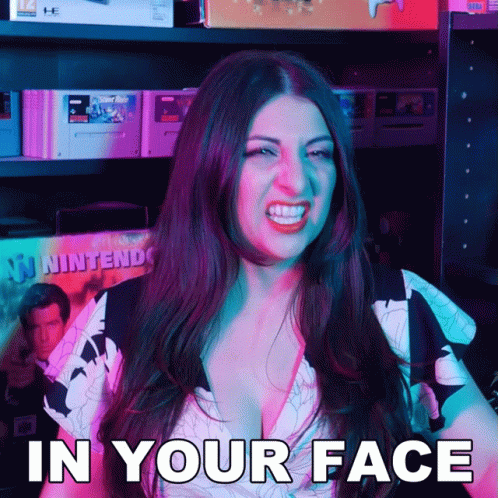 A Woman Says In Your Face While Standing In Front Of A Shelf Full Of Video Games GIF