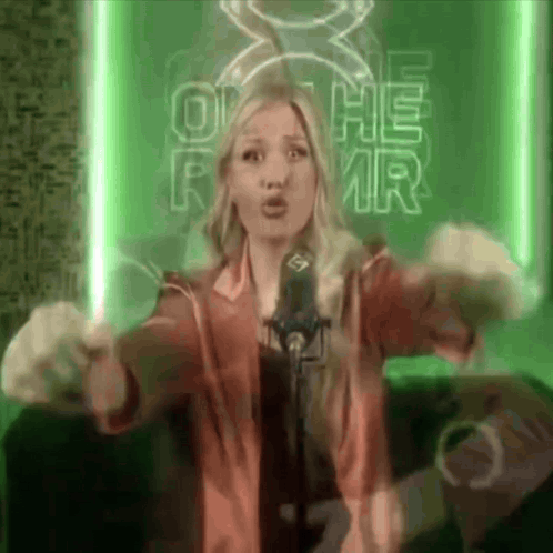 A Woman Singing Into A Microphone In Front Of A Sign That Says O He Rmr GIF