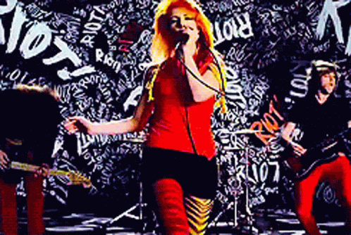 A Woman Singing Into A Microphone In Front Of A Wall With The Word Riot Written On It GIF