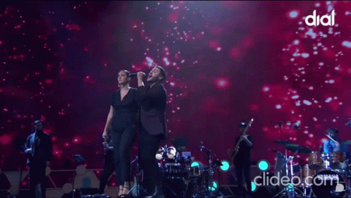 A Woman Singing Into A Microphone Next To A Man On A Stage With Dial Written On The Bottom GIF