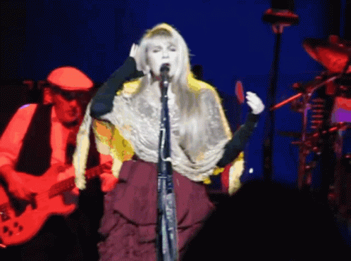 A Woman Singing Into A Microphone While A Man Plays A Guitar In The Background GIF