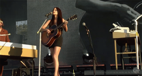 A Woman Singing Into A Microphone While Holding A Guitar On Stage GIF
