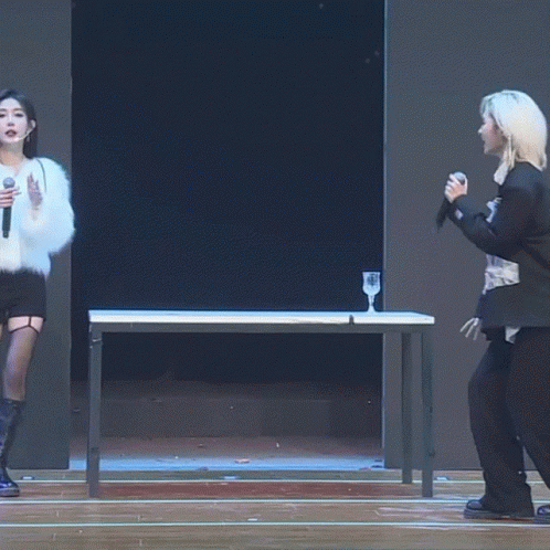 A Woman Singing Into A Microphone While Standing Next To A Table GIF