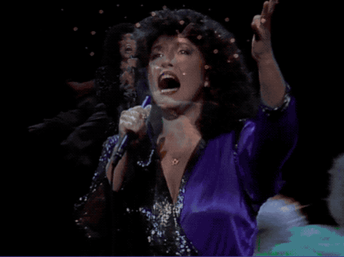 A Woman Singing Into A Microphone While Wearing A Purple Shirt GIF