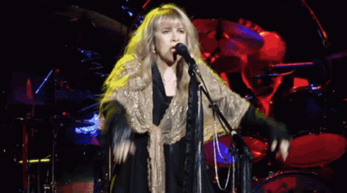 A Woman Singing Into A Microphone With A Drum Set In The Background GIF