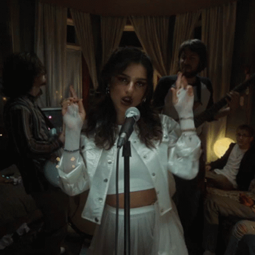 A Woman Singing Into A Microphone With A Man Playing A Guitar Behind Her GIF
