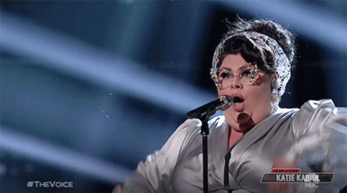 A Woman Singing Into A Microphone With A Sign That Says Katie Kabuk On It GIF