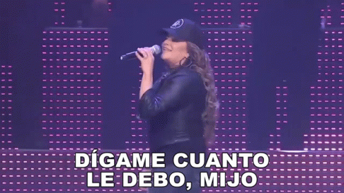 A Woman Singing Into A Microphone With The Words " Digame Cuanto Le Debo Mijo " Behind Her GIF