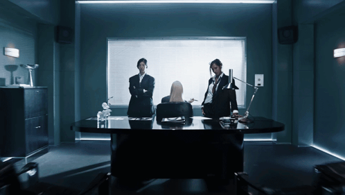 A Woman Sits At A Desk In A Dark Room With Two Men Standing Around Her GIF
